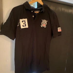 Svart pikétröja Vinson Polo Club - Svart pikétröja från Vinson Polo Club med klassisk krage, korta ärmar och broderad logga på bröstet. Snygg patch med siffran 3 på ena sidan och amerikansk flagga på ärmen. Perfekt för dig som gillar sportig och stilren look.