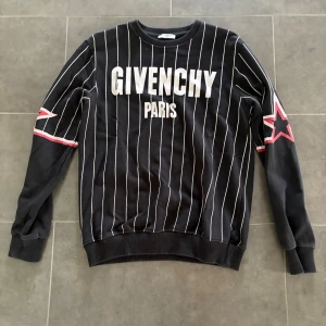 Svart randig Givenchy sweatshirt - Svart sweatshirt från Givenchy med vita vertikala ränder och stor logga på bröstet. Röda och vita stjärndetaljer på ärmarna ger en sportig vibe. Mjuk bomullskänsla och ribbade muddar vid ärmslut och midja. Perfekt för dig som gillar streetstyle.