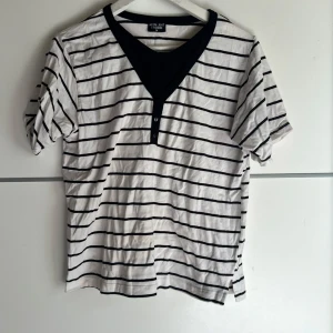 Randig t-shirt från Moon Light Fashion - Vit och svart randig t-shirt från Moon Light Fashion i storlek M. T-shirten har korta ärmar, svart v-ringad kant med knappdetalj framtill och är gjord i mjuk bomull. Perfekt för en chill och stilren look.