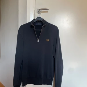 Svart half zip tröja från Fred Perry - Mörkblå/ svart långärmad tröja från Fred Perry med half zip och guldfärgad logga på bröstet. Ribbstickade muddar med vita och gula ränder vid ärmslut. Klassisk och stilren design, perfekt för lager-på-lager. Materialet är mjuk bomull. Två små limm fläckar kar som ej går bort enligt mig som syns på bilderna. Storlek xs som passar som xs också. Skriv om ni har frågor!