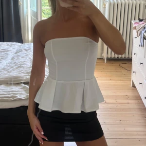 Vit bandeau topp med volang från H&M - Supersnygg vit bandeau topp från H&M med volang nertill. Toppen är axelbandslös och har en figurnära passform upptill som går ut i en peplumvolang. Perfekt att styla med kjol eller jeans för en trendig look.