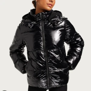 Glansig svart pufferjacka med huva - Snygg svart pufferjacka med glansig finish och dragkedja framtill. Helt jätte bra skick utan någon defekter. Passformen är normal och jackan är lätt att matcha med olika outfits.