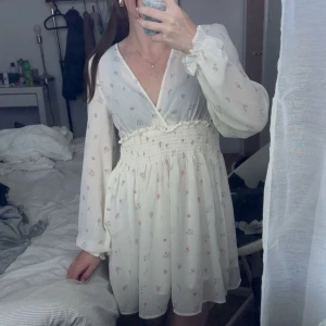 Vit blommig omlottklänning XS - Supersöt vit kortklänning med små blommor i pastellfärger, omlott över bysten och smockad midja. Långa, luftiga ärmar med resår vid handlederna. Perfekt för vår och sommar, ger en romantisk och fräsch vibe.