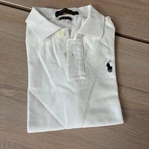 Vit pikétröja från Ralph Lauren M - Klassisk vit pikétröja från Ralph Lauren. Tröjan har korta ärmar, krage och knäppning framtill med två knappar. Ikoniska broderade loggan i mörkblått på bröstet. Tillverkad i bomull för en skön och luftig känsla.