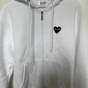 Vit hoodie Comme des Garçons Play - Vit hoodie från Comme des Garçons Play med dragkedja, huva och snörning. Ikoniskt svart hjärta med ögon broderat på bröstet. Mjuk bomullskvalitet och känguruficka framtill. Perfekt för en clean och stilren look. Tar även byten 