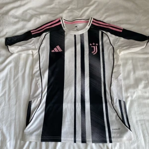 Juventus randig fotbollströja Adidas - Juventus fotbollströja från Adidas med klassiska svarta och vita ränder, rosa detaljer på axlar och logga. Kortärmad modell i lätt och ventilerande Aeroready-material. Perfekt för dig som vill sticka ut på planen eller visa ditt stöd för Juve. Kan passa på folk som har storlek M också.
