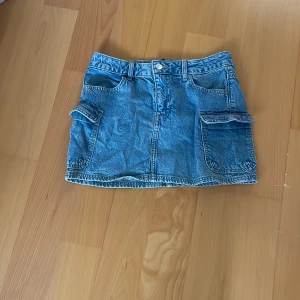 Blå jeanskjol med stora fickor - Snygg blå jeanskjol i kort modell med stora fickor på sidorna och bälteshällor. Klassisk denimlook som passar perfekt till sneakers eller boots. Kjolen har knapp och dragkedja framtill och coola detaljer som ger en avslappnad vibe.