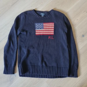 Mörkblå stickad tröja Ralph Lauren - Mörkblå stickad tröja från Ralph Lauren med amerikansk flagga och RL-broderi på bröstet. Klassisk rund halsringning och ribbade muddar. Perfekt för dig som gillar preppy stil och vill sticka ut lite extra. Storleken ör 18-20 så den passar s/m