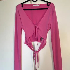 Rosa omlottopp med knyt från NA-KD - Trendig rosa omlottopp från NA-KD med långa ärmar och snygga knytband både framtill och baktill. Toppen har v-ringning och croppad passform som ger en cool och modern look. Perfekt för dig som vill sticka ut med färg och stil.
