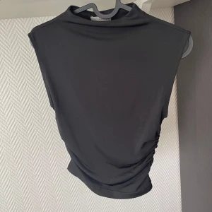 Svart ärmlös polotopp från H&M - Snygg svart polotopp från H&M med hög hals och ärmlös design. Toppen har en tight passform och cool rynkdetalj på ena sidan som ger en modern look. 