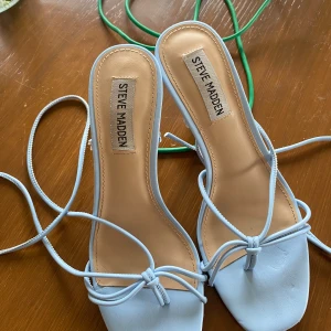 Ljusblå klackar från Steve Madden - Snygga ljusblå klackar  från Steve Madden med tunna remmar och knytning runt vristen. Skorna har fyrkantig tå och låg klack, tillverkade i syntetmaterial med en stilren och modern look. Perfekta för att lyfta din outfit med en pop av färg. Skorna har slitningar som ni ser på bilderna 