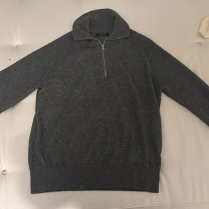 Grå stickad half zip-tröja Massimo Dutti - Mörkgrå stickad tröja från Massimo Dutti med hög krage och half zip-dragkedja. Tröjan har långärmade ribbade ärmar och en klassisk, lite loose passform. Perfekt för kyliga dagar och enkel att matcha med jeans eller chinos.
