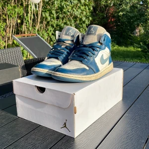 Nike Air Jordan 1 Mid blå/vit - Nike Air Jordan 1 Mid i blått och vitt skinn med klassisk Jordan-logga på sidan och tungan. Skorna har blå snörning, perforerad tå och vit mellansula. Snygg retrostil med högt skaft och rund tå, perfekt för dig som gillar streetwear och sneakerskultur. Tyvär lite förstörd inuti ena skon men går och ha på sig. 