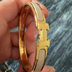 Harmens Clic H armband guld & beige - Snyggt Harmens Clic H armband i guldmetall med beige emalj och ikoniskt H-spänne. Stilren och lyxig accessoar som lyfter vilken outfit som helst. Perfekt för dig som gillar exklusiva detaljer och klassisk design.
