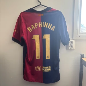 FC Barcelona Raphinha matchtröja Nike M - Säljer en officiell FC Barcelona fotbollströja från Nike i storlek M, med Raphinha och nummer 11 på ryggen. Tröjan har klassiska blå och röda paneler, guldiga detaljer och klubbmärke på bröstet. Tillverkad i lätt och ventilerande material, perfekt för fotbollsälskare.