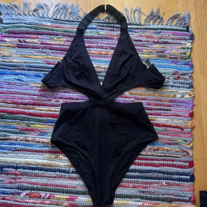 Svart cut-out bikini med spänne - Snygg svart bikini med cut-out design och djup v-ringning. Halterneck med breda band och silvrigt spänne framtill. Hög midja och stilren look, perfekt för stranden eller poolen. Materialet känns mjukt och följsamt.