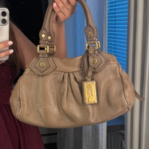 Marc by Marc Jacobs Väska  - Säljer min super snygga trendiga väska från Marc by Marc Jacobs!! Modellen kallas för ”classic q baby groovy” det är alltså den mindre storleken. Väskan har en super fin beige/brun färg med snygga guldetaljer. Passar verkligen till allt😍 Den är i mycket bra skick, lite smått sliten på vissa ställen men absolut inget man tänker mycket på. Väldigt svår att få tag på därav priset, Har inte axelbandet så det får man inte med tyvärr. Skriv vid frågor/intresse, kom gärna med bud! 💞🙌