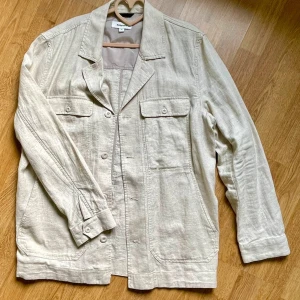 Beige overshirt från Kappahl i linne - Snygg beige overshirt från Kappahl i linne. Jackan har klassisk krage, knäppning framtill, två bröstfickor med lock och knappar samt två sidfickor. Perfekt för lager-på-lager och chill stil. Lätt och luftig känsla.