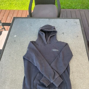 Mörkblå Peak Performance hoodie - Snygg svart hoodie från Peak Performance med klassisk logga på bröstet. Tröjan har huva, känguruficka och ribbade muddar vid ärmslut och nederkant. Perfekt för chill dagar eller när du vill ha en enkel och stilren look.