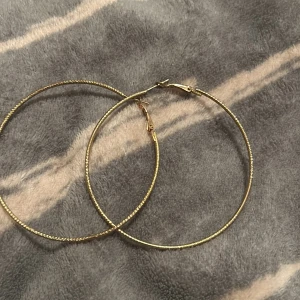 Stora guldiga hoops örhängen - Säljer ett par stora, tunna hoops i guldton. Örhängena har en enkel men snygg design med lätt tvinnad yta som ger lite extra glans. Perfekta för dig som gillar statement-smycken och vill ha något som syns.