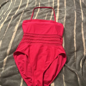 Rosa baddräkt med smala axelband - Snygg rosa baddräkt med smala, justerbara axelband och dekorativa sömmar över magen. Enkel och stilren design utan mönster, med vadderade kupor för extra stöd. Perfekt för strand eller poolhäng i sommar.