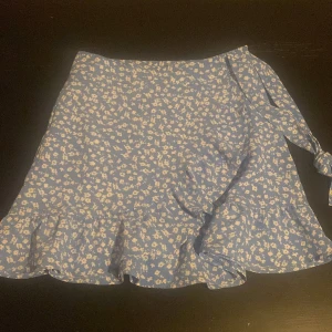 Blommig blå omlottkjol från FB Sister - Supersöt blå kortkjol från FB Sister med vitt blommönster. Kjolen har omlottmodell med knytband i sidan och volang längst ner för extra flowig känsla. Perfekt för dig som gillar en feminin och trendig stil.