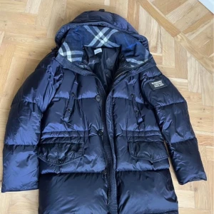 Mörkblå dunjacka från Burberry - Snygg mörkblå dunjacka från Burberry med klassiskt rutigt foder i luvan och tydlig logga på ärmen. Jackan har stora fickor framtill, dragkedja och knappar. Perfekt för kalla dagar och riktigt stilren look. En av knapparna har en liten bit som knäckts inget som stör eller syns direkt.