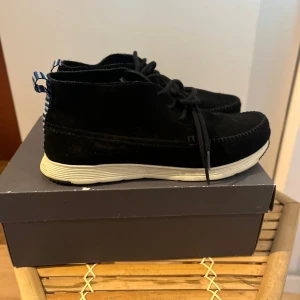 Svarta mockasneakers från Ransom - Svarta sneakers från Ransom i mjuk mocka med vita, räfflade sulor och blåvitrandiga dragflikar bak. Skorna har snörning och en stilren, rundad form som passar till många outfits. Perfekta för dig som gillar en clean och modern look.