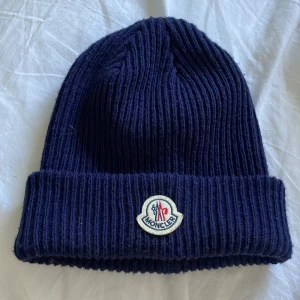 Mörkblå Moncler mössa - Snygg mörkblå stickad mössa från Moncler med klassisk uppvikt kant och broderad logotyp framtill. Perfekt accessoar för kyliga dagar och enkel att matcha med olika outfits. 
