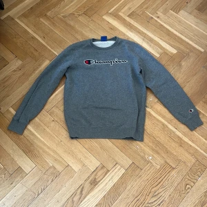 Grå Champion sweatshirt X-Large - Grå sweatshirt från Champion i storlek X-Large. Klassisk crewneck med broderad Champion-logga på bröstet och liten logga på ärmen. Mjuk insida och ribbade muddar vid ärmslut och midja. Perfekt för en chill och sportig stil.