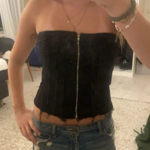 Svart korsett-topp från Zara - Säljer en svart korsett-topp från Zara i storlek XS. Toppen är axelbandslös och har en dragkedja framtill samt råa kanter nertill för en edgy vibe. Snygg passform som sitter tight och markerar midjan. Perfekt att styla med jeans eller kjol.