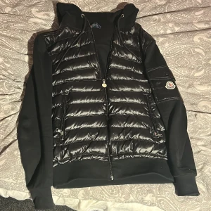 Moncler cardigan - Svart cardigan från Moncler med glansig quiltad front, huva och dragkedja. Ärmar och rygg i mjuk bomull, med Moncler-logga på ena ärmen. Snygg streetstil. Använd cirka 5 gånger. Inga fel på den utan en repa på ena knappen, alla scans funkar på den och kommer direkt till monclers hemsida, skriv vid funderingar eller ge prisförslag 