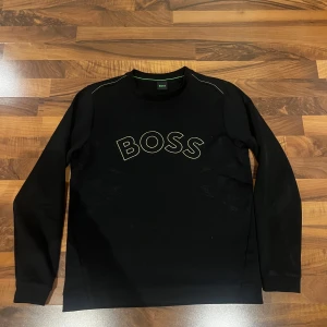 Hugo Boss Sweatshirt  - Svart sweatshirt från BOSS med broderad logga i guld på bröstet. Tröjan har rund halsringning, långa ärmar och en clean, stilren look. Perfekt för dig som gillar klassiska streetwear-vibbar och vill sticka ut med en snygg logga.