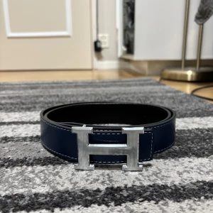 H bälte - Snyggt mörkblått skärp med ikoniskt H-spänne i silverfärgad metall. Bältet har vändbar design med svart baksida, perfekt för att lyfta vilken outfit som helst.