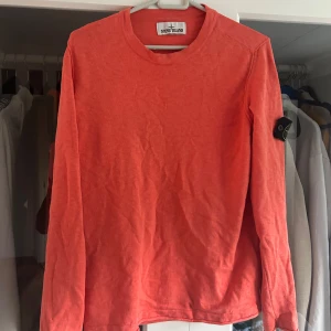 Korallröd långärmad Stone Island tröja - Snygg korallröd långärmad tröja från Stone Island med klassisk rund hals och ikonisk logga på vänster ärm. Tröjan är i mjuk bomull och har en clean, enkel look som passar perfekt till jeans eller cargobyxor.