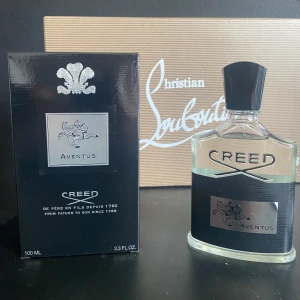 Creed Aventus 100ml Eau de Parfum - Lyxig parfym från Creed, Aventus 100ml. Flaskan är transparent med svart etikett och silverdetaljer, samt svart kork. Kommer med originalkartong och tillbehör. Doften är ikonisk och exklusiv, perfekt för dig som vill sticka ut med en elegant och modern signatur.