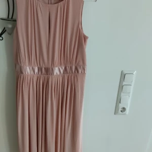 Ljusrosa knälångklänning från Bodyflirt - Elegant ljusrosa långklänning från Bodyflirt med ärmlös design och vackra veck framtill. Klänningen har ett glansigt band i midjan och en liten slits vid halsringningen. Perfekt för dig som vill ha en stilren och feminin look.