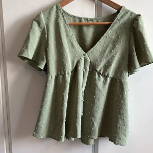 Ljusgrön blus med knappar och v-ringning - Supersöt ljusgrön blus med v-ringning och knappar framtill. Blusen har korta, vida ärmar och ett  prickigt strukturmönster i tyget. Vet ej vart den är i från eller storlek men skulle gissa på XS/S. 