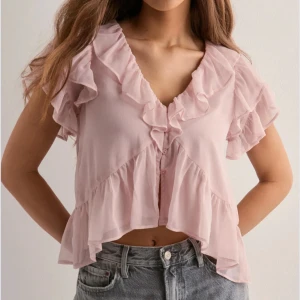 Ljusrosa volangblus med knappar - Supersöt ljusrosa blus med volanger längs v-ringningen och ärmarna. Blusen är croppad och har knappar framtill, luftigt material. Slutsåld på hemsidan 🩷