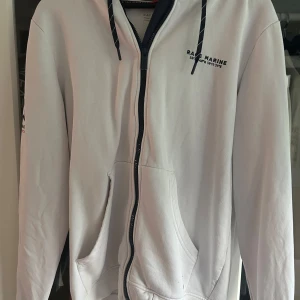 Vit hoodie från Race Marine med dragkedja - Vit hoodie från Race Marine med svart dragkedja, huva med snörning och broderad logga samt siffran 83 på ärmen. Stora fickor framtill och marinblå detaljer. Perfekt för dig som gillar sportig och clean stil.