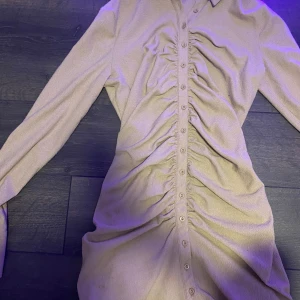 Beige skjortklänning från Bik Bok - Snygg beige skjortklänning från Bik Bok med långa ärmar och knappar hela vägen framtill. Klänningen har cool rynkdetalj längs mitten som ger en unik look. Perfekt för dig som gillar stilrena men trendiga plagg. använd endast en gång