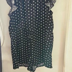 Svart prickig byxdress Shein Curve - Svart byxdress från Shein Curve med vita prickar, volangdetaljer vid ärmhålen och dragkedja bak. Klänningen har en lös passform och är tillverkad i ett lätt polyestertyg, perfekt för dig som gillar en lekfull och trendig stil.