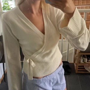Vit omlottkofta från Gina Tricot - Superfin vit omlottkofta från Gina Tricot med knyt i sidan och breda ribbade muddar. Koftan har lång ärm och är i mjukt stickat material, perfekt för lager på lager. Snygg v-ringning och klassisk passform.