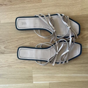 Beige sandaler med remmar från Zara - Stilrena sandaler från Zara i beige med tunna remmar och fyrkantig tå. Platt sula och minimalistisk design, perfekta för varma dagar. Snygga detaljer med knytning framtill och svarta kanter runt sulan.