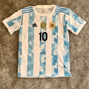 Argentina Messi 10 Adidas fotbollströja - Säljer en officiell Argentina landslagströja från Adidas med Messi och nummer 10 på ryggen. Tröjan har klassiska blå och vita ränder, broderade märken och Copa America 2021-märke. Tillverkad i lätt och ventilerande Aeroready-material, perfekt för fotbollsfans.
