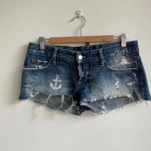 Dsquared slitna jeansshorts med ankare - Riktigt coola blå jeansshorts från Dsquared med slitna detaljer, fransig kant och broderat ankare framtill. Klassisk femficksmodell med Dsquared-logga på bakfickan. Perfekta för sommaren och ger en edgy vibe till din outfit.