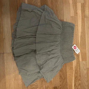 Grå volangkjol med smockmidja - Supersöt grön kortkjol med volanger och bred smock i midjan. Kjolen har en lätt rutig struktur och är perfekt för dig som gillar en luftig och trendig stil. Passar till sneakers eller boots för en chill look.