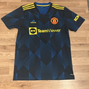 Manchester United bortatröja Adidas - Snygg Manchester United bortatröja från Adidas med blått och svart geometriskt mönster, gula detaljer och klubbmärke på bröstet. Kortärmad modell i lätt och ventilerande material, perfekt för match eller träning. TeamViewer-logga på framsidan och klassiska Adidas-ränder på axlarna.