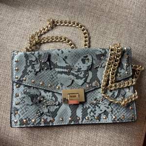 Grå axelväska med kedja från Mango - Snygg väska men guld detaljer. Perfekt storlek och kedjan går att justera. Ormskinnsmönster men lite fjärilar och liknande på framsidan. I fint skick men de är lite slitage på spännet (sista bilden) 