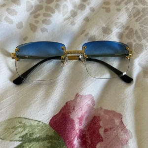 Blå-guld solglasögon från Lumière - Snygga solglasögon från Lumière med blåtonade glas och gulddetaljer. Halvramlös design med svarta skalmar och elegant guldmönstrad brygga. Kommer med originalfodral, putsduk och förvaringspåse. Perfekt accessoar för en cool look.